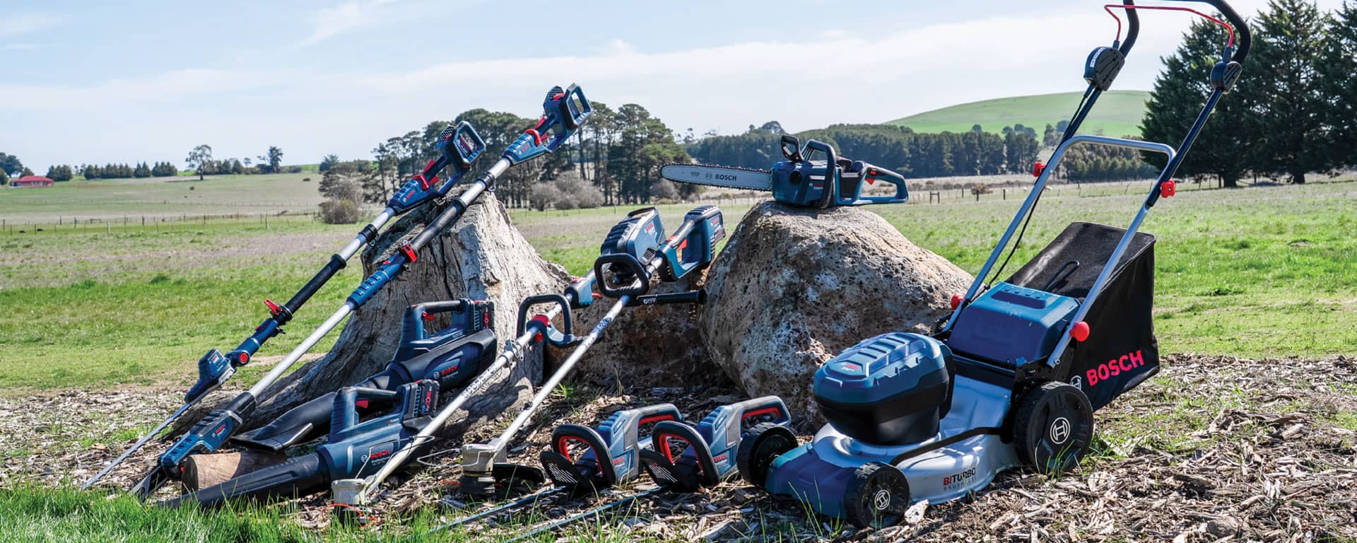 A collection of Bosch gardening tools on a tree stump in a green landscape.