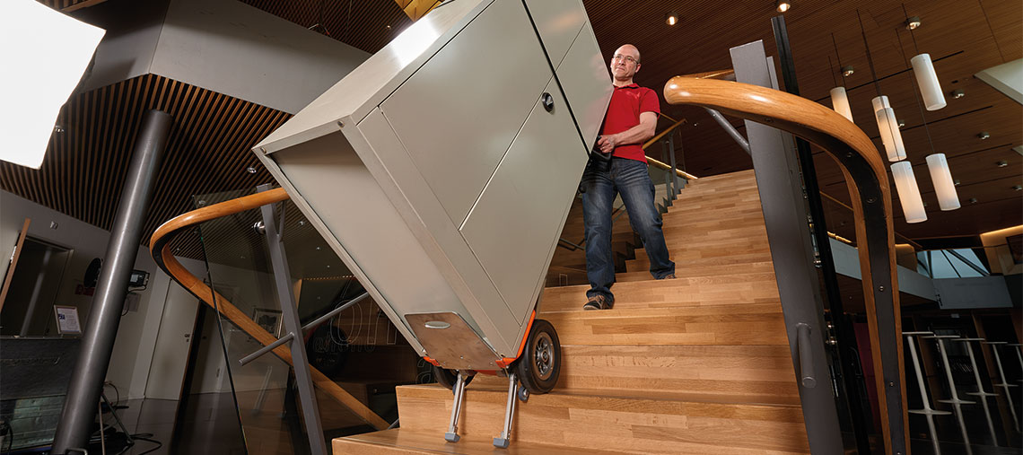 A man is carrying a silver cabinet up a wooden staircase.