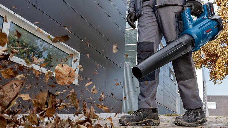 A man with a blue Bosch leaf blower is blowing leaves in front of a building.