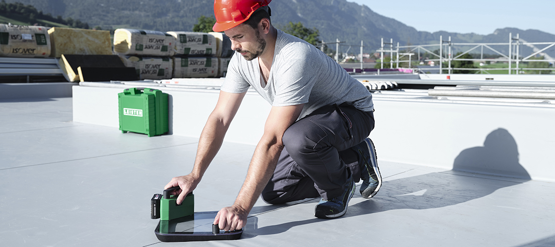 A man in a red helmet kneels on a light gray roof and measures with a laser device.