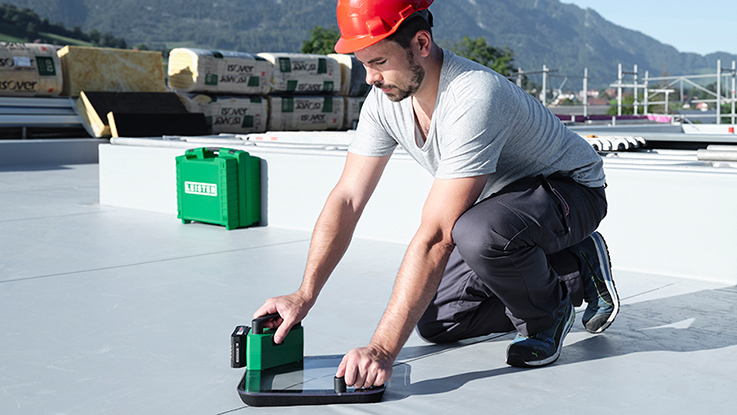 A craftsman in a gray T-shirt and red helmet kneels on a flat roof. He uses a green tool and a black ruler to take precise measurements. In the background, building materials like insulated rolls are visible, and the mountains are slightly blurred. The sky is blue and sunny, and the scene appears focused and professional.