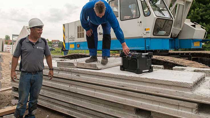 Two construction workers stand by a crane; one operates a device on concrete slabs.