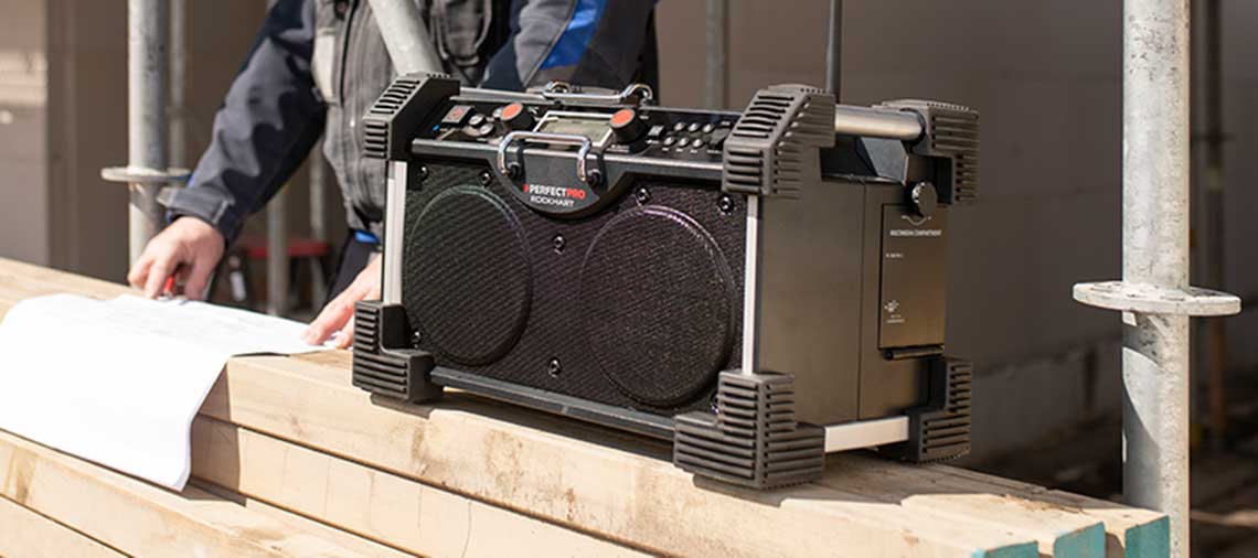 A black PerfectPro construction radio is standing on a wooden board.