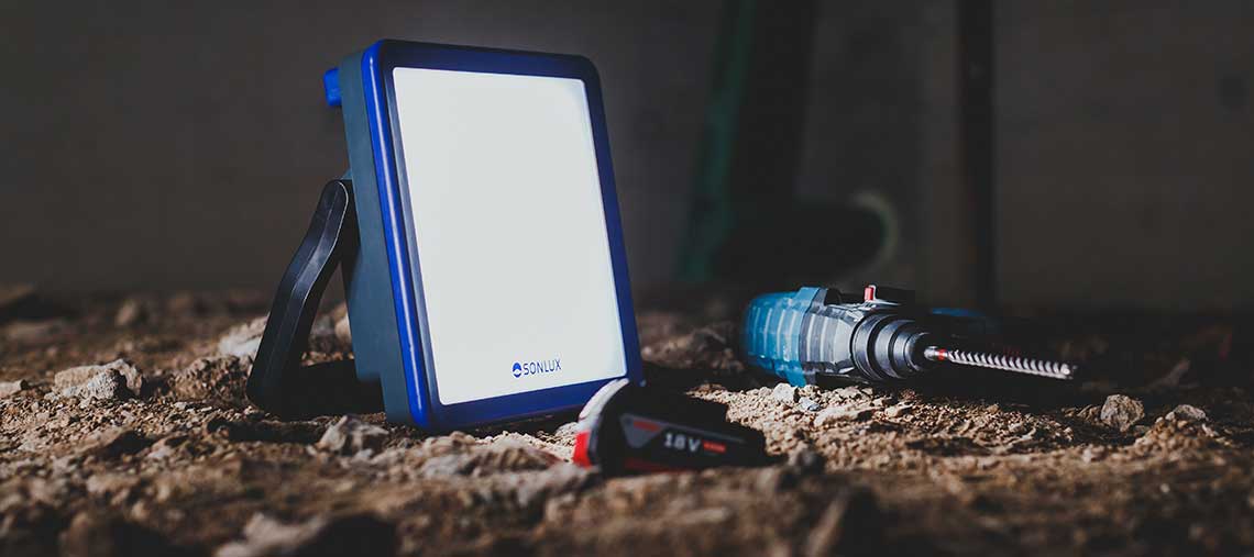 A blue SONLUX construction lamp is standing on the floor next to a battery drill.