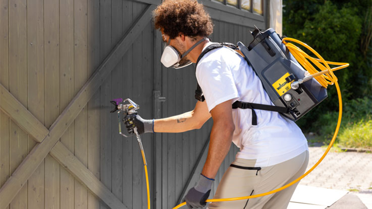 A man is spraying paint on a gray wooden door with a spray device.
