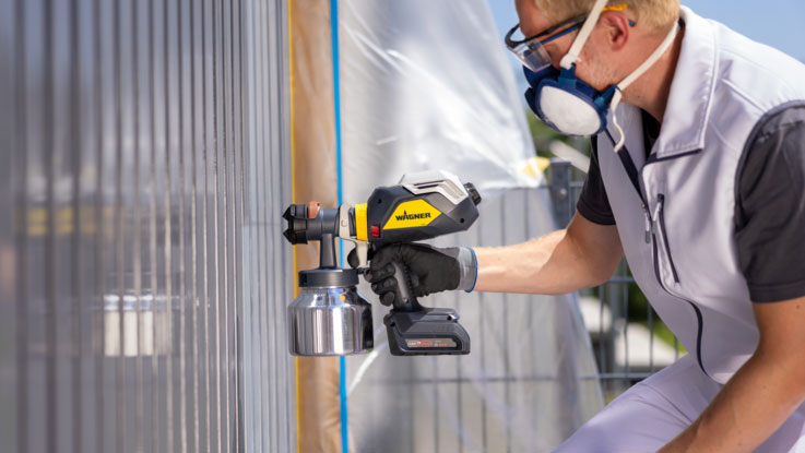 A craftsman is spraying paint with a Wagner spray gun onto a wall.