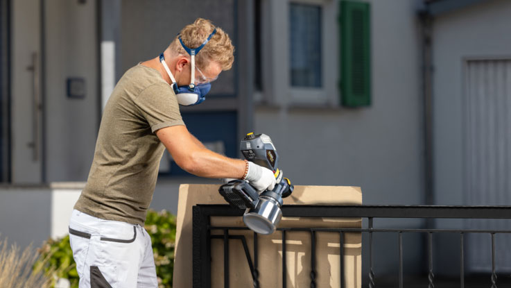 A man with a mask sprays paint with a Bosch tool on a wall.