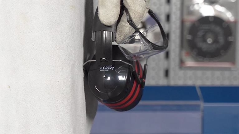 Safety goggles and black ear protectors with red stripes hang on a wall.