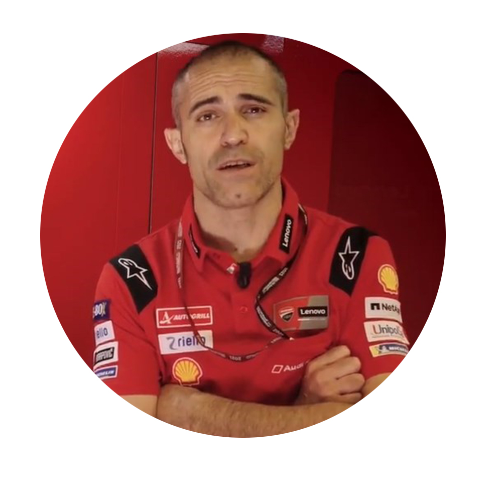 A man in a red shirt stands in front of a red background and speaks.