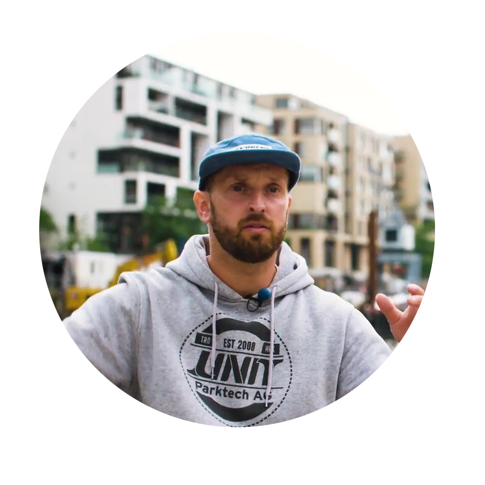 A man in a gray hoodie and blue hat speaks in front of modern buildings.