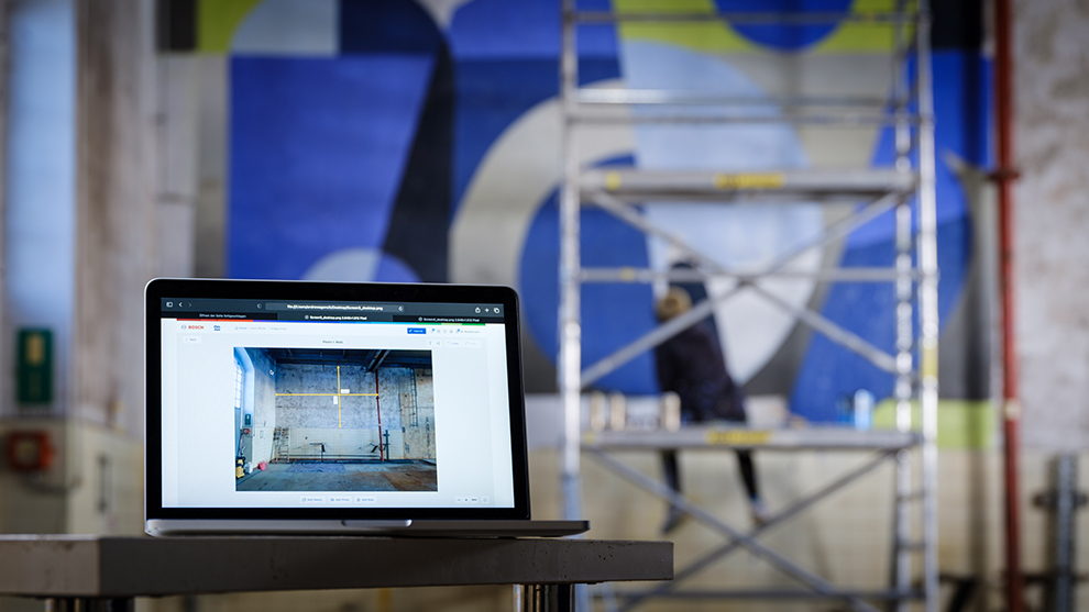 Laptop shows an image of a gray wall, scaffolding in the background, a person painting.