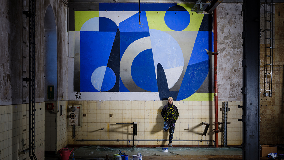 Boy in a colorful hoodie stands in front of a geometric mural in an empty room.