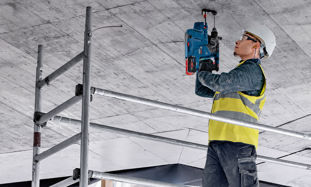 A worker in a yellow vest is using a blue Bosch drill on the ceiling.