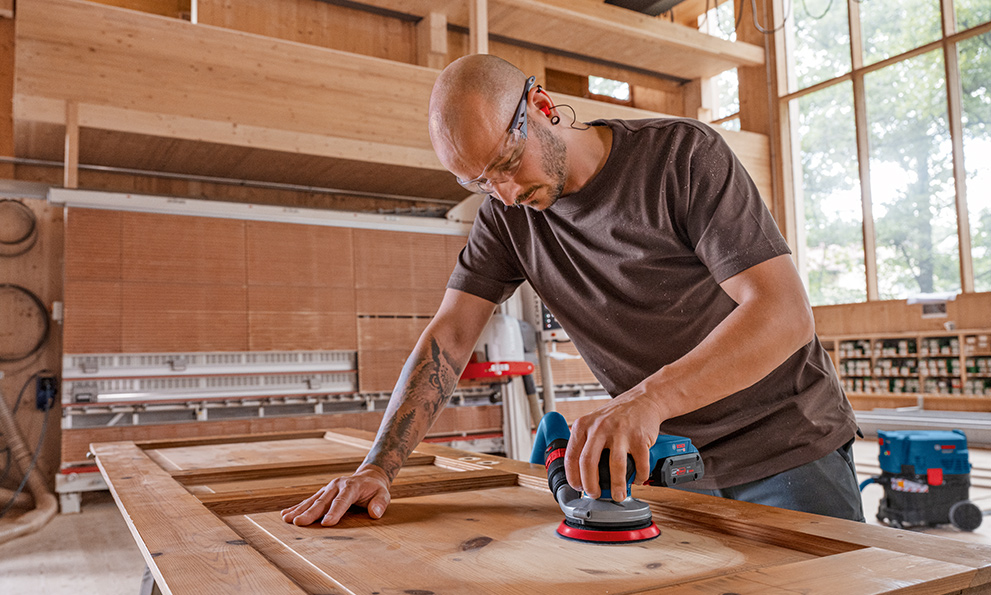 Sanding a door A man is sanding wood at a table in a bright workshop.