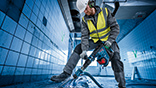 A worker in a yellow vest is using a Bosch hammer drill in a room.