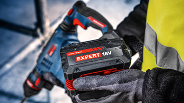 A worker holds a black battery with red lettering "EXPERT 18V" in his hand.