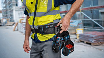 A worker in a yellow safety vest stands on a construction site and reaches for tools in his belt pocket.