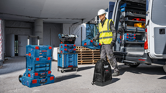 A worker in a safety vest and helmet stands next to a van, surrounded by blue Bosch Professional toolboxes.