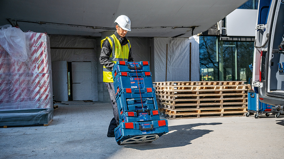 A worker in a yellow safety vest and helmet lifts a blue Bosch Professional tool case at a construction site. In the background, wooden pallets and construction accessories are visible.