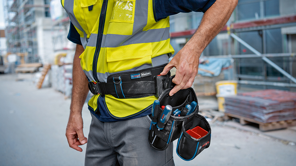 A craftsman in a yellow safety vest reaches into his tool bag to take out tools. Construction materials are visible in the background.
