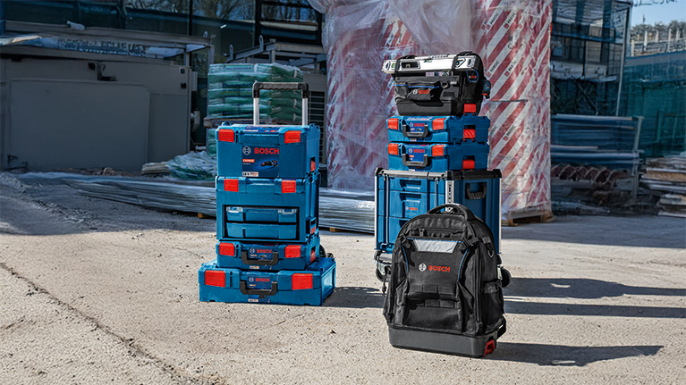 Several stackable blue Bosch Professional toolboxes are on a construction site floor next to a black backpack.