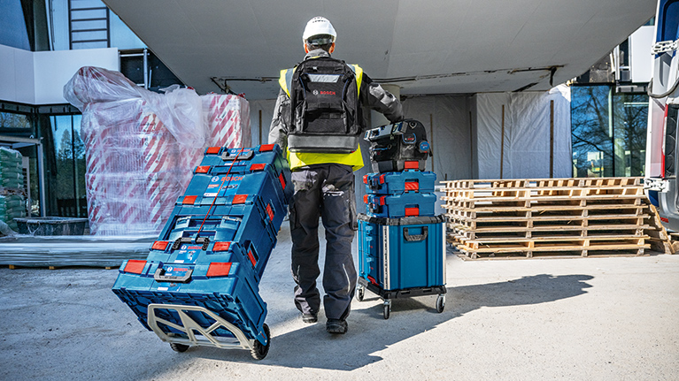 A worker in safety clothing is pushing two blue toolboxes from Bosch Professional on a construction site.