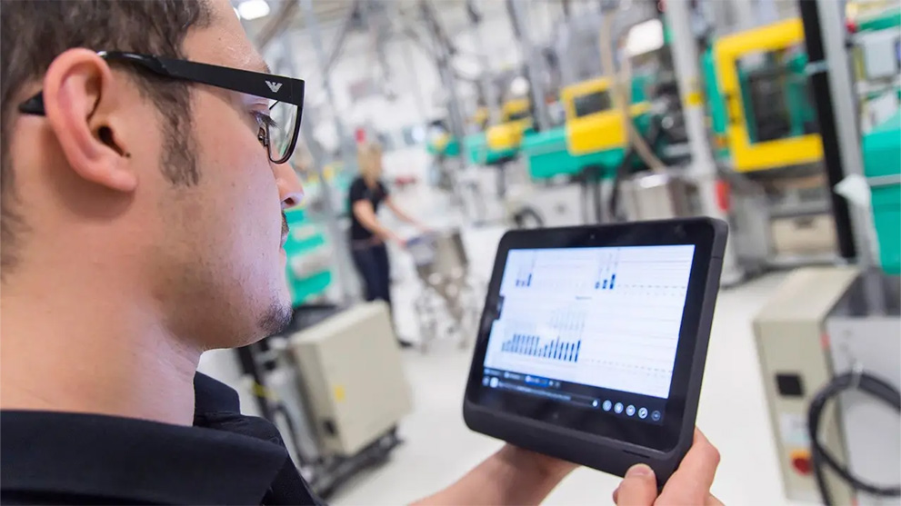 A man with glasses is looking at a tablet in a factory hall.