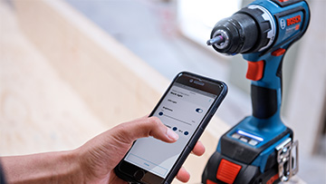 A hand operates a smartphone next to a blue Bosch Professional cordless screwdriver.