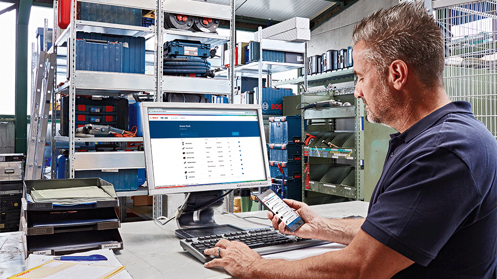 Man at computer with Bosch tools in the background, using smartphone.