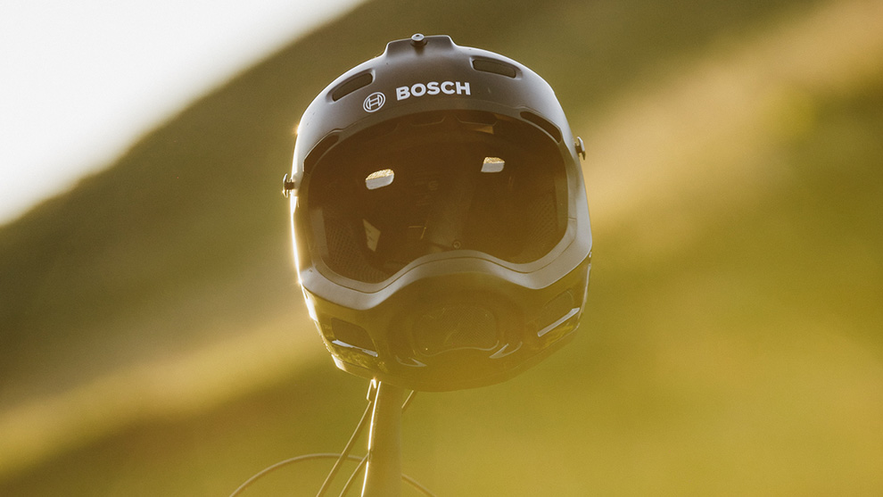 A black Bosch helmet hangs in front of a blurred, sunny landscape.