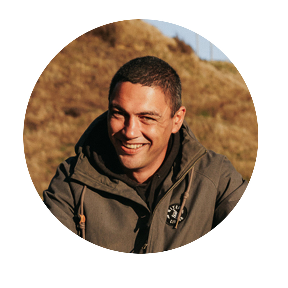 A laughing man in a gray parka sits in front of a meadow.