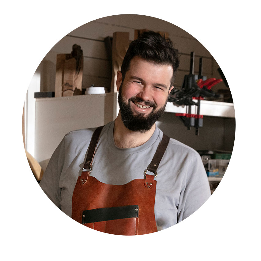 A smiling man in a leather apron stands in a workshop.