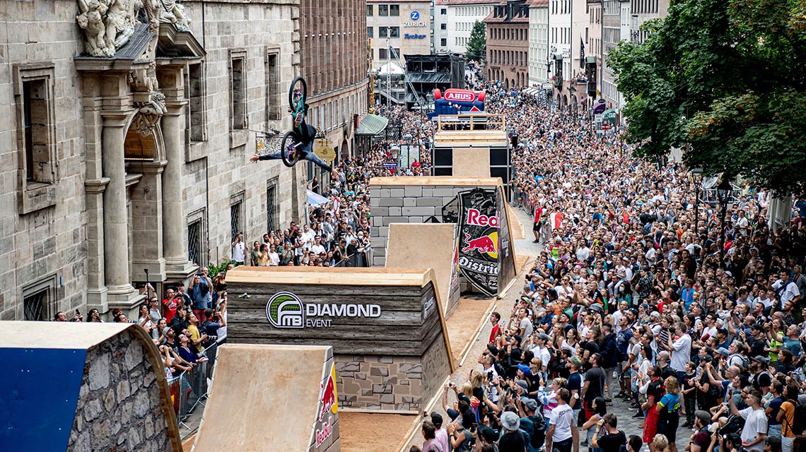 A BMX rider jumps over ramps in front of a large crowd.
