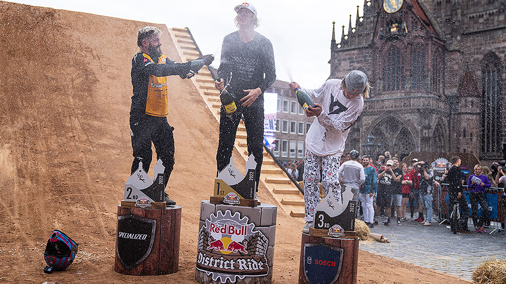 The champions of Red Bull District Ride 2022 pop bottles of champagne on the podium Three winners celebrate on podiums with champagne, a crowd in the background.