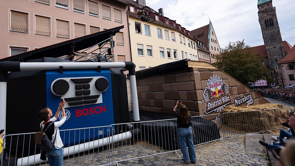 A rider performs a trick in the air after flying off the gigantic Bosch Up Box  Bosch advertising setup with spectators, stunt bike, and historic building in the background.