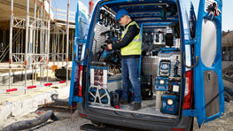 A tradesman in a safety vest is working in the open trunk of a blue van.