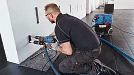A craftsman kneels next to a wall and uses a Bosch drill.